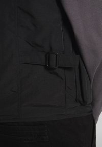Black padded jacket featuring a belt and buckle detail, textured fabric, and layered design with a grey sweatshirt underneath.
