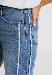 Denimjeans med en frynset sidesøm, lyseblå vask, design med fem lommer og kobbernitter, der viser en slank pasform set fra siden på en model.
