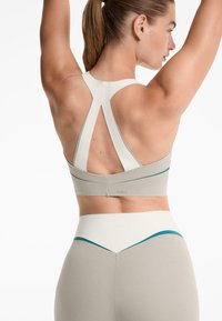 Kvinna med upplyfta armar, klädd i en beige och vit sport-BH samt högmidjade leggings med teal-accenter, som vänder sig bort mot en vit bakgrund.