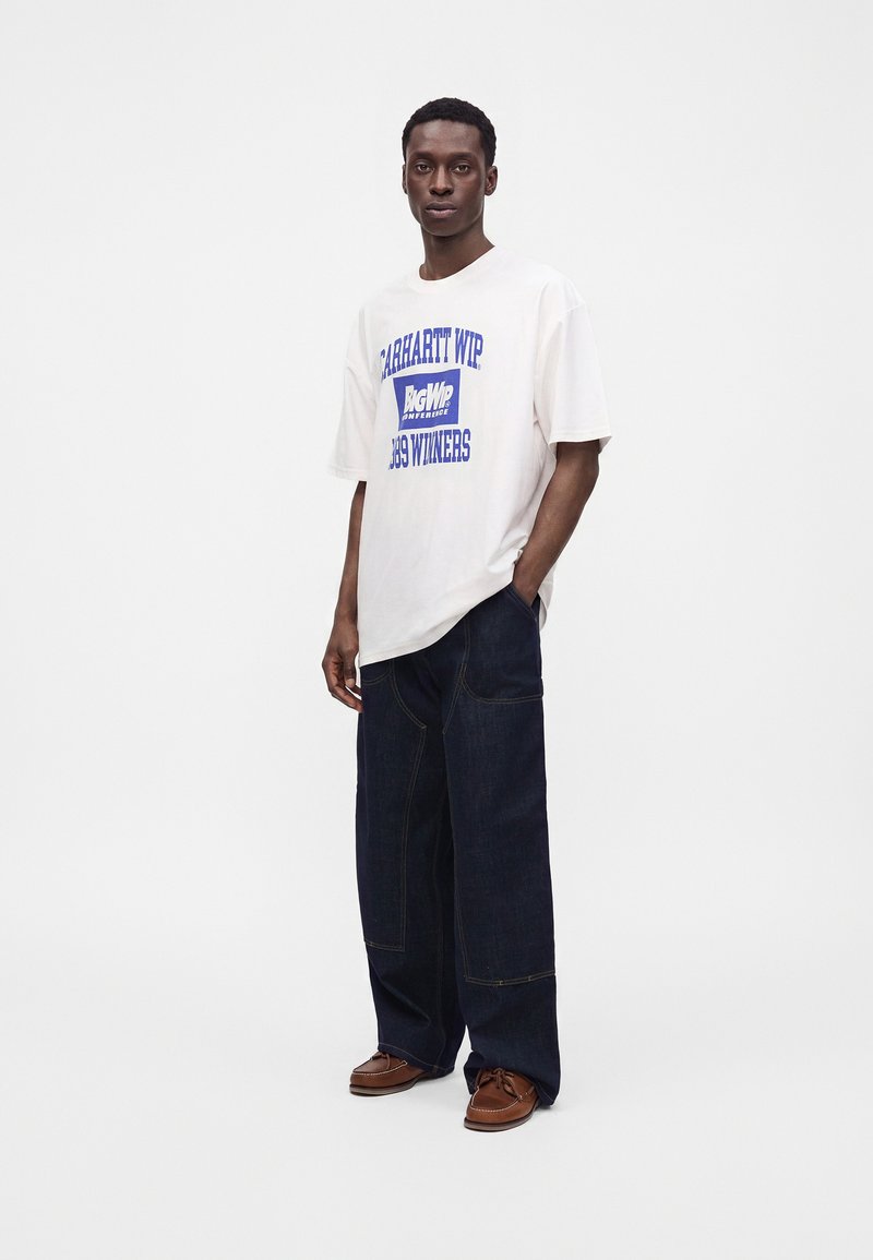 Man wearing white graphic T-shirt, wide dark blue jeans, and brown shoes standing with one hand in pocket against plain white background.