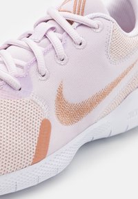 Zapatilla deportiva de color púrpura claro con material de malla, que presenta un logo y acentos en color melocotón-naranja de Nike, punta redondeada y suela texturizada.