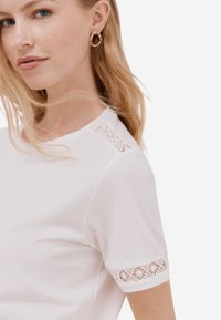 Bílé tričko s krátkým rukávem a květinovou krajkovou vencí na manžetě. Měkká tkanina, crew neckline, přiléhavý design, minimalistický styl.