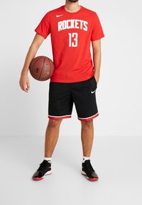 Czerwona koszulka Nike Rockets z białymi napisami, czarne siatkowe spodenki z czerwonymi akcentami i czarne sportowe buty z czerwonymi detalami. Trzyma piłkę do koszykówki.