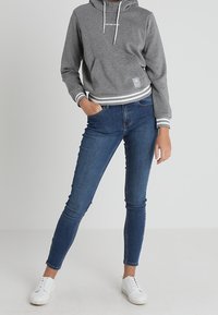Sudadera gris recortada con cordones blancos y puños a rayas, combinada con jeans skinny azul oscuro y zapatillas blancas. Diseño simple, look casual.