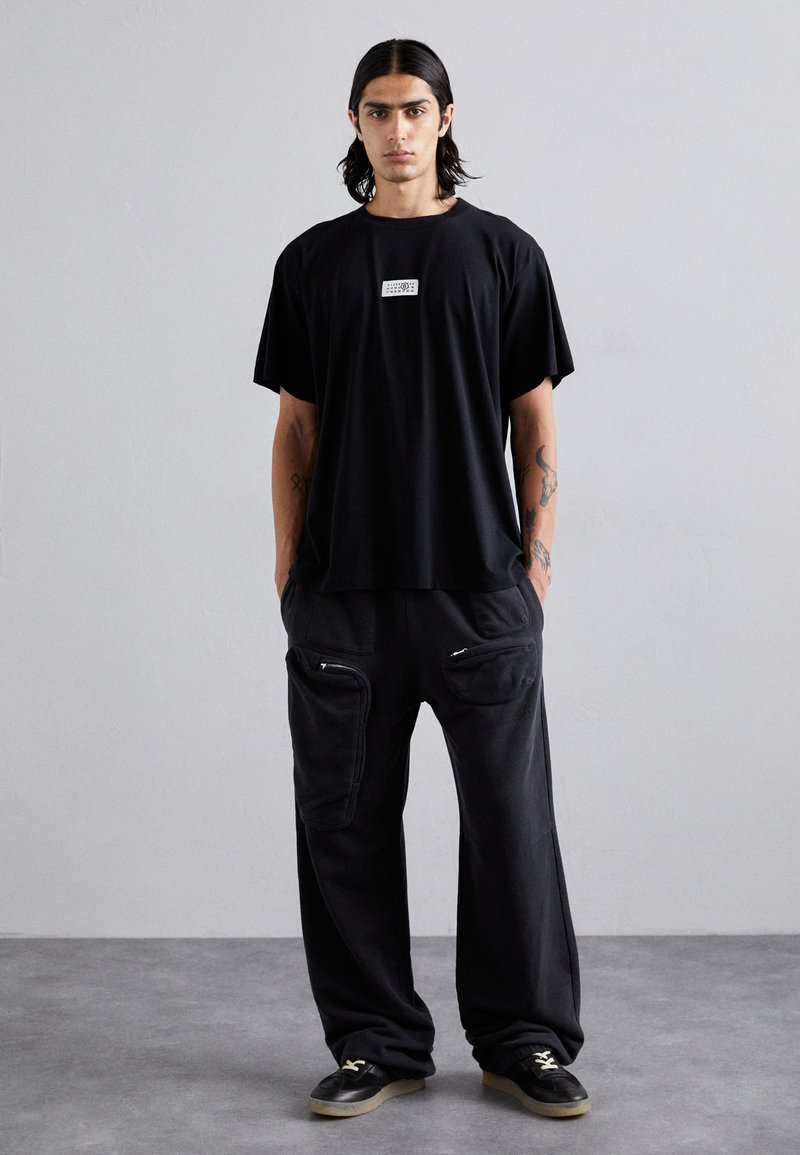 T-shirt nera oversize con un logo grafico bianco sul petto, abbinata a pantaloni neri larghi con tasche laterali zipperate.