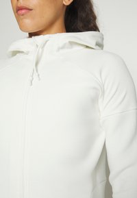 Vit zip-up hoodie i strukturerad tyg, med huva och dragsko samt raglanärmar för en figurnära silhuett.