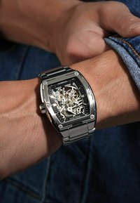 Zegarek automatyczny o szkieletowym designie, czarna tarcza, srebrna stal nierdzewna, prostokątna koperta oraz widoczne mechanizmy i tryby.