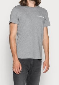 Šedé bavlněné tričko s kulatým výstřihem, krátkými rukávy a bílým logem "Calvin Klein Jeans" na levé straně hrudníku. Měkká textura, uvolněný střih.