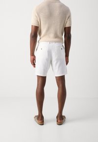 Pantaloni corti bianchi in lino con due tasche posteriori e bottoni marroni. Indossati con una maglietta in maglia testurizzata beige e sandali marroni.