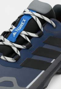 Μπλε αθλητικό παπούτσι με μαύρες λεπτομέρειες, υφασμένο πάνω μέρος, κορδόνια με λευκό και μαύρο σχέδιο, και γλώσσα σε γκρι χρώμα με μπλε λογότυπο.