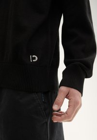 Schwarzer Strickpullover mit geripptem Saum und Bündchen, mit einem kleinen weißen Logo an der unteren Vorderseite. Hand, die nahe an schwarzen Hosen ruht.