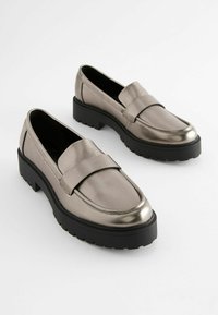 Mocasines plateados metálicos sin cordones con una textura suave, puntera redonda y suela de goma negra gruesa con un fondo acanalado.