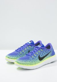 Une paire de chaussures de course Nike bleues et vert néon avec le logo swoosh noir et des semelles blanches, posée sur une surface blanche.
