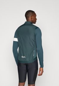 Tumši zaļš velosipēdu veste ar augstu apkakli, viegls materiāls, elastīga apakšmala, atstarojoši balti akcenti un "Rapha" logotips aizmugurē.