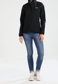 Veste softshell noire avec un col montant, une fermeture éclair sur le devant, des poches latérales et un petit logo sur le côté gauche de la poitrine. Portée avec un jean bleu et des baskets.