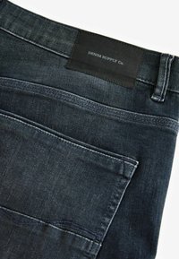 Donker denim jeans met een leren label met "DENIM SUPPLY Co." en een gestikt zakdetail. Gladde textuur met een fitted ontwerp.