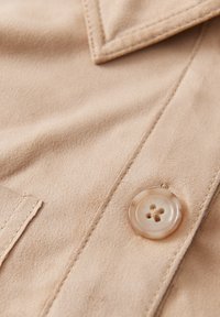 Beige suède stof met een gladde textuur, voorzien van een duidelijke knop met vier gaten en een puntige kraag. Gedetailleerde stiksels op de randen.