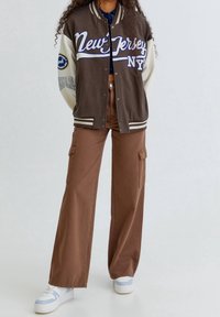 Brązowa kurtka varsity z kremowymi rękawami, białymi literami i naszywkami. Połączona z szerokimi brązowymi spodniami cargo i białymi sneakersami.