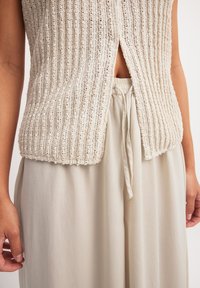Gilet en tricot beige avec texture côtelée, présentant une fente à l'avant ; associé à un pantalon beige léger et fluide accentué par un cordon de serrage.