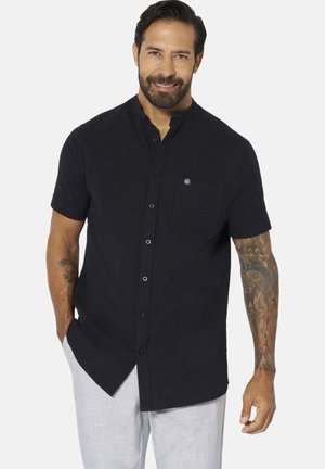 Man with beard wearing a black short-sleeve button-up shirt and light gray pants, smiling with tattoos visible on his right forearm.