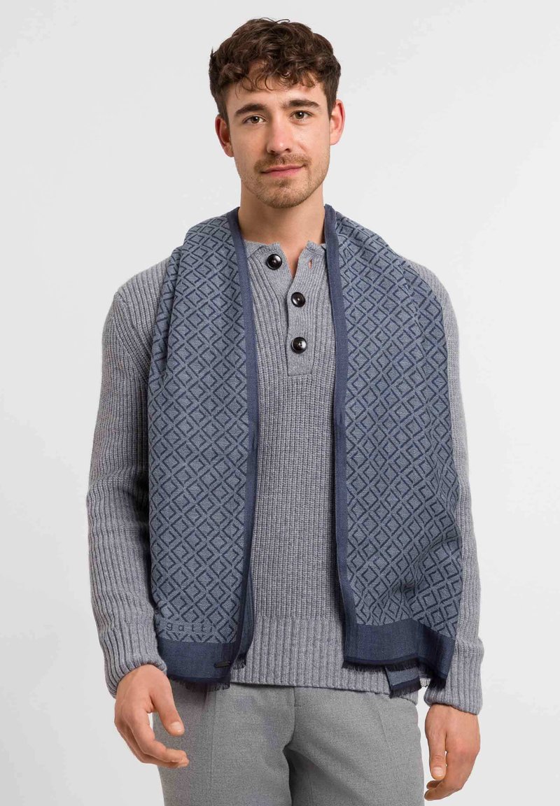 Bufanda de estampado marino con diseño geométrico, superpuesta a un suéter ribeteado gris claro con cuello henley y botones oscuros.