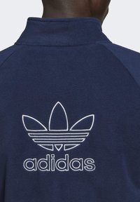 Mörkblå fleecejacka med hög krage och broderad vit Adidas trefoil-logga på ryggen, med en mjuk textur.