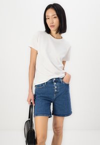 Hvit bomullsskjorte med korte ermer, paired med høyt liv blå denimshorts med knappestolpe og rå kanter. Svart håndveske holdes i hånden.