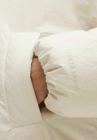 Svetlo béžová púzdrová bunda s naťahovacími rukávmi, s jemne embosovaným logom. Tkanina má hladkú, mierne zvrásnenú štruktúru.