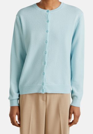 Personne portant un cardigan bleu clair boutonné à manches longues, associé à un pantalon beige taille haute, sur un fond uni.