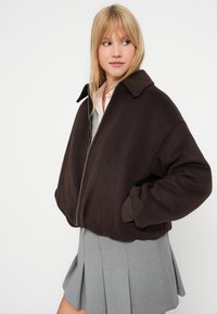 Brązowa, wełniana kurtka o kroju cropped z dużym kołnierzem, zapinana na zamek, o luźnym kroju, w zestawieniu ze szarym, plisowanym spódnicą.