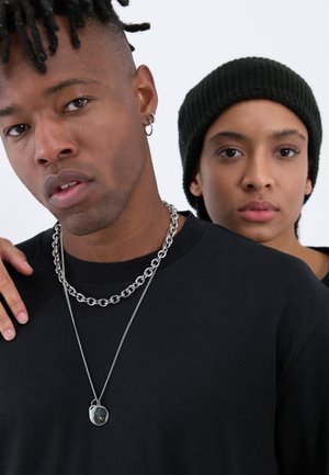 Black T-shirt worn by a person with short, textured hair and chain necklaces. A second person in a black beanie stands closely behind.