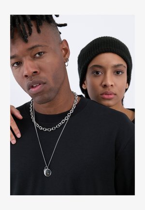 Svart T-skjorte brukt av en person med kort, teksturert hår og kjedehalskjeder. En annen person i en svart beanie står tett bak.