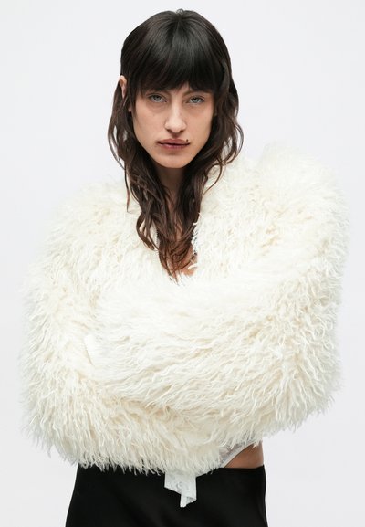 Λευκό πέπλο από αφράτη γούνα με μακριές υφές και χαλαρή εφαρμογή, σε στρογγυλεμένο σχήμα. Φοριέται πάνω από σκοτεινή φούστα για αντίθεση.