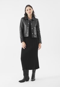 Schwarze Lederjacke in cropped Länge mit zwei Fronttaschen, kombiniert mit einem langen schwarzen Rippkleid und schwarzen Cowboystiefeln, vor einem neutralen Hintergrund.