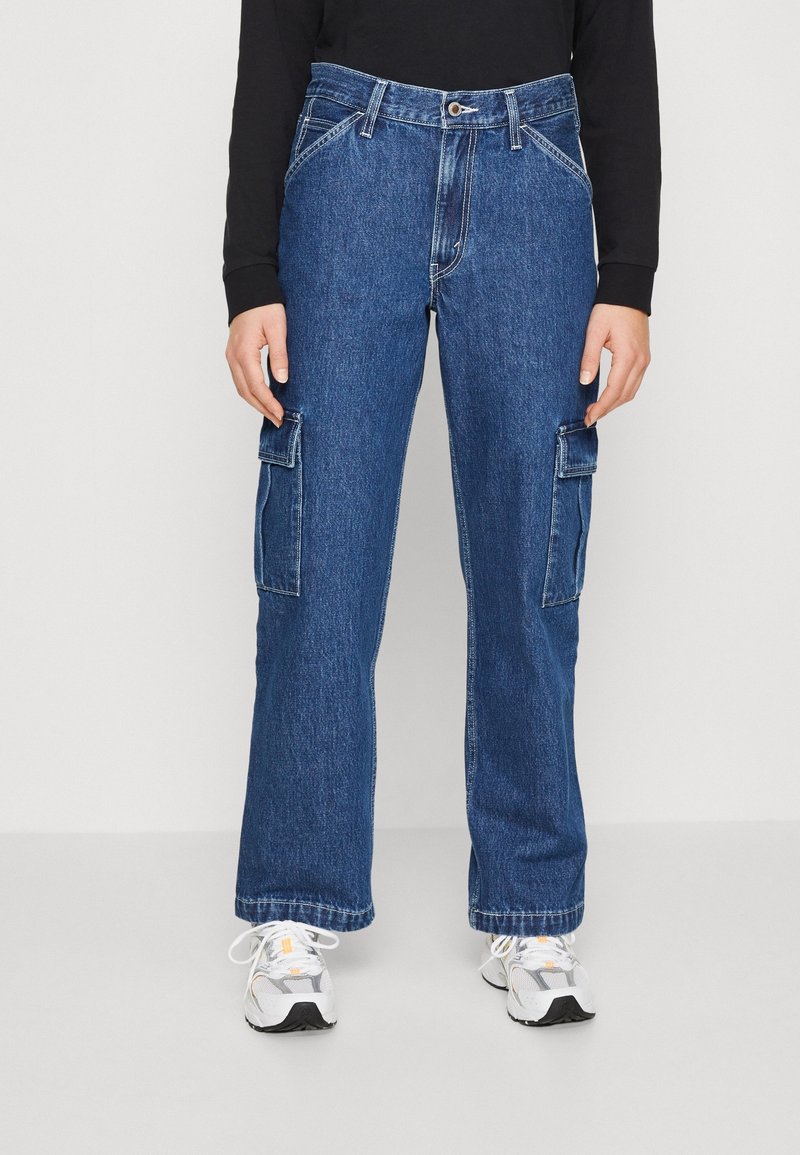 Pantalon cargo en denim bleu à coupe décontractée, doté de deux poches latérales, d'une fermeture à bouton et de détails de couture apparents. Assorti à des baskets blanches.