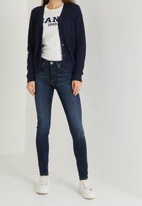 Cárdigan de punto azul marino, camisa blanca con texto en azul marino, jeans oscuros y zapatillas blancas. El cárdigan cuenta con un diseño de botones en la parte delantera y puños acanalados.