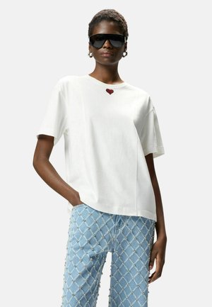 Mujer con una camiseta blanca de gran tamaño con un pequeño detalle de corazón rojo, vaqueros azul claro con tachuelas, grandes gafas de sol negras y pendientes de aro.