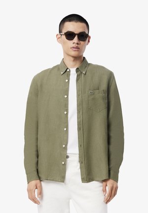 Camisa de lino verde oliva de botones con un bolsillo en el pecho y botones blancos, usada sobre una camiseta blanca. El modelo lleva gafas de sol.