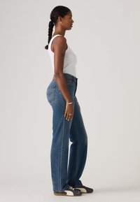 Τζιν παντελόνι από μπλε ντένιμ με ψηλή μέση και άνετη εφαρμογή, με πίσω τσέπες και κλείσιμο με κουμπί, φορεμένο με λευκό αμάνικο τοπ και αθλητικά παπούτσια.