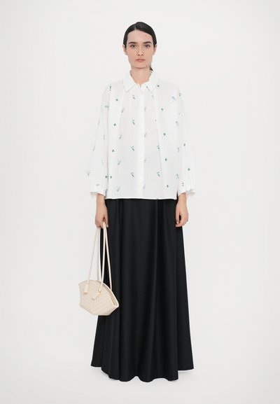 Chemise blanche avec des motifs floraux verts, associée à une longue jupe noire. Un sac à main en osier beige complète la tenue.