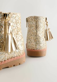 Botas doradas brillantes con una superficie texturizada, que cuentan con un cierre lateral y borlas decorativas, y una suela sólida en color rosa-marrón.