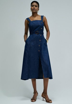 Femme portant une robe midi en denim bleu sans manches avec boutons et ceinture, debout les mains dans les poches avec des sandales marron à talons.