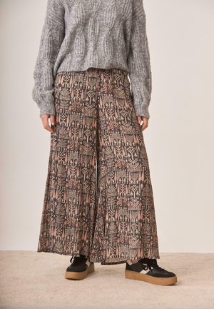 Persona con pantalones anchos estampados, suéter gris de punto texturizado y zapatillas negras con suelas gruesas color beige sobre alfombra beige.