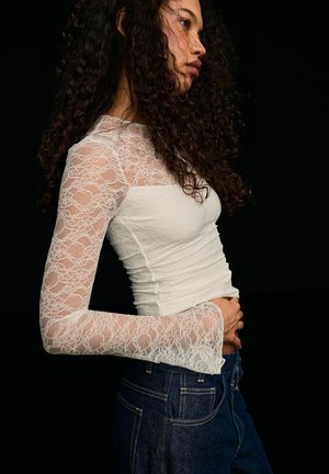 Femme aux cheveux bouclés portant un haut en dentelle blanche à manches longues et un jean bleu foncé, posant de profil sur un fond noir.