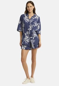 Una mujer está de pie luciendo una camisa floral azul y pantalones cortos a juego, con zapatillas blancas, sonriendo con confianza frente a un fondo sencillo.