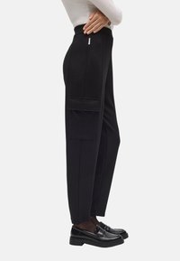 Pantalones de tela negra de cintura alta con corte recto, bolsillos laterales y textura de tela suave. Combinados con mocasines negros brillantes.