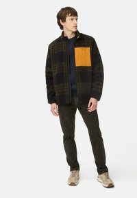 Fleecejacke mit einem schwarz-grünen Karomuster und einer leuchtend orangefarbenen Brusttasche. Kombiniert mit dunklen Hosen und beigen Sneakers.