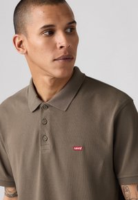 Bruine polo van gestructureerde stof met een kraag en drie knopen. Bevat een klein rood Levi's-logo op de linkerkant van de borst.