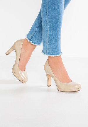 Kvinde iført lyseblå jeans med flossede kanter og beige skinnende højhælede sko, stående på en hvid gulv.