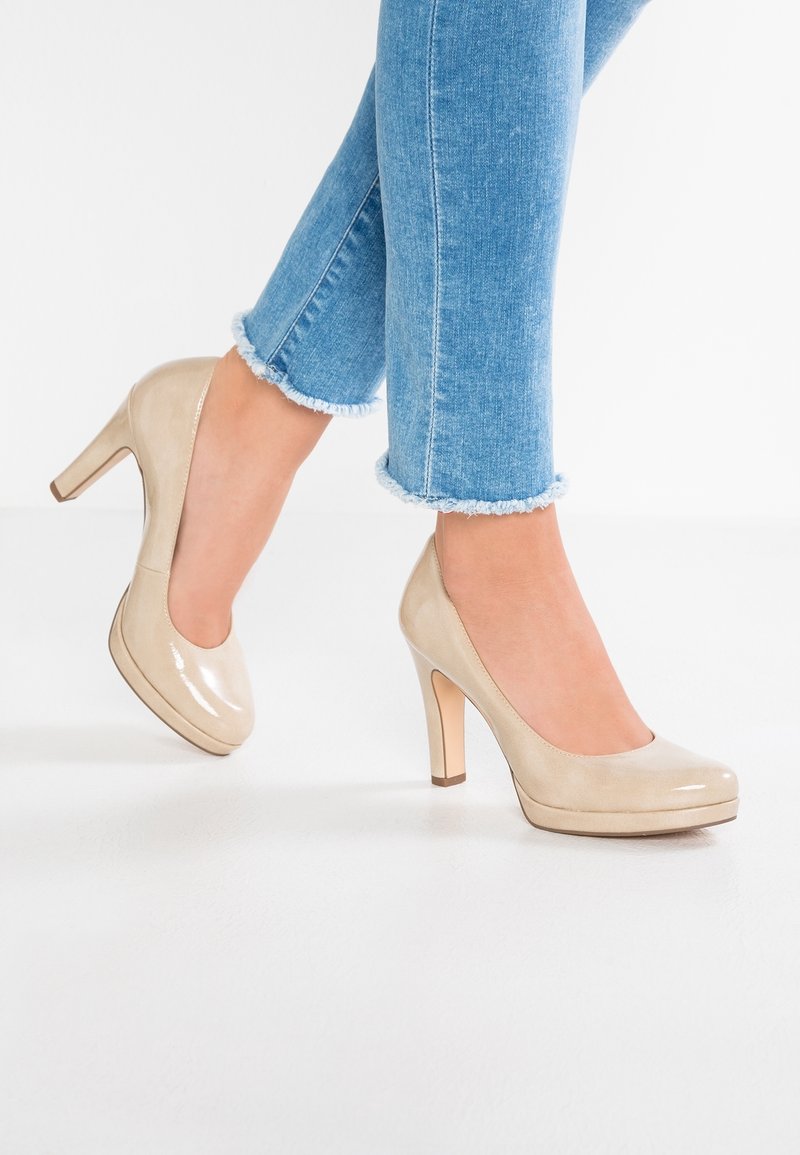 Femme portant un jean bleu clair à ourlet effiloché et des chaussures à talons hauts beige brillantes, debout sur un sol blanc.